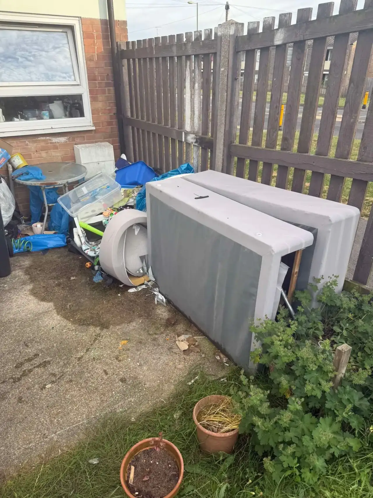 A mattress and divan base propped up outside beside a fence